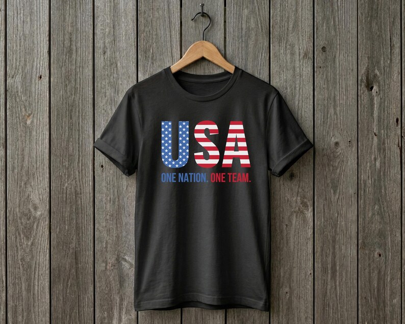 Peut inclure: Un t-shirt noir &agrave; manches courtes avec le texte USA dans un motif de drapeau am&eacute;ricain rouge, blanc et bleu. Sous les grandes lettres, le texte indique ONE NATION. ONE TEAM. en bleu et rouge. Le t-shirt est sur un cintre en bois.