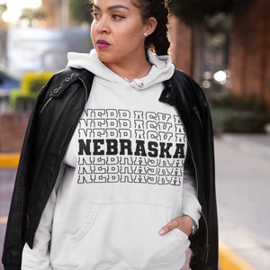 Op de afbeelding: Witte hoodie met zwarte tekst die meerdere keren "NEBRASKA" herhaalt in een gestapelde vorm. Het model draagt een zwart leren jack over de hoodie.