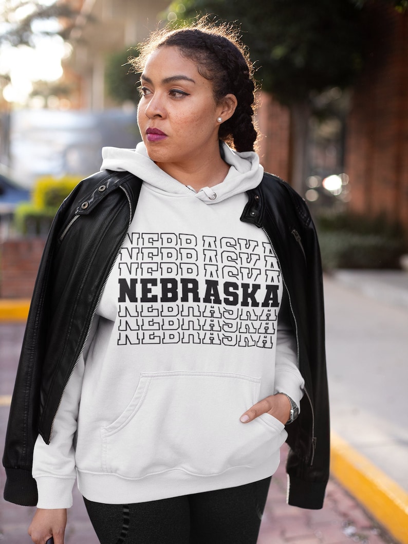 Op de afbeelding: Witte hoodie met zwarte tekst die meerdere keren "NEBRASKA" herhaalt in een gestapelde vorm. Het model draagt een zwart leren jack over de hoodie.
