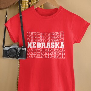 Op de afbeelding: Rood T-shirt met een witte grafische print van het woord "NEBRASKA" dat vijf keer in een gestapelde vorm wordt herhaald.