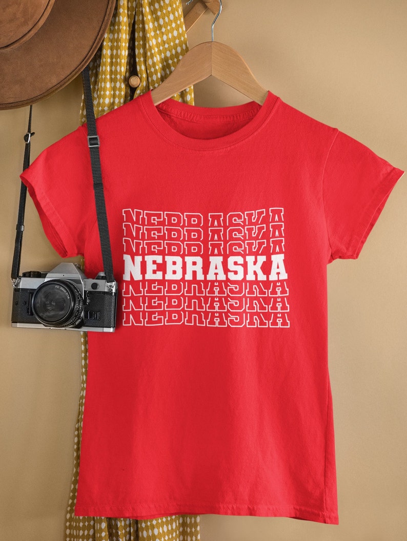 Op de afbeelding: Rood T-shirt met een witte grafische print van het woord "NEBRASKA" dat vijf keer in een gestapelde vorm wordt herhaald.