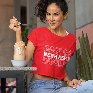 Op de afbeelding: Een vrouw draagt een rood, gecropped T-shirt met de tekst "NEBRASKA" die in het wit wordt herhaald. Ze draagt een blauwe jeans en heeft donker haar.
