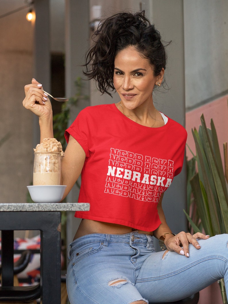 Op de afbeelding: Een vrouw draagt een rood, gecropped T-shirt met de tekst "NEBRASKA" die in het wit wordt herhaald. Ze draagt een blauwe jeans en heeft donker haar.