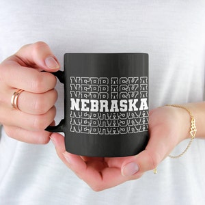 Op de afbeelding: Zwarte koffiemok met witte tekst die "NEBRASKA" herhaalt in een retro stijl.