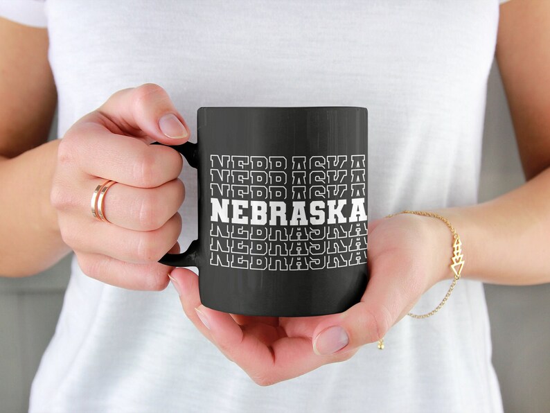 Op de afbeelding: Zwarte koffiemok met witte tekst die "NEBRASKA" herhaalt in een retro stijl.