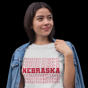 Op de afbeelding: Een wit T-shirt met een rode grafische print van het woord "NEBRASKA" dat meerdere keren in een retro-stijl wordt herhaald.