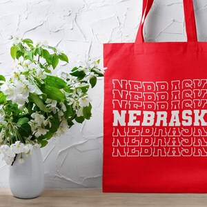 Op de afbeelding: Rode tote bag met witte tekst die meerdere keren "NEBRASKA" zegt. De tas staat voor een witte muur met een boeket witte bloemen in een witte vaas.