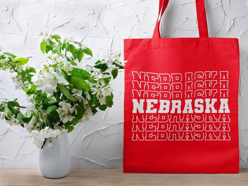 Op de afbeelding: Rode tote bag met witte tekst die meerdere keren "NEBRASKA" zegt. De tas staat voor een witte muur met een boeket witte bloemen in een witte vaas.