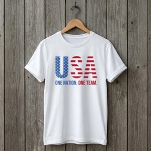 Peut inclure: Un t-shirt blanc &agrave; manches courtes avec le texte USA dans un motif bleu &eacute;toil&eacute; et ray&eacute; rouge et blanc. Sous les lettres, le texte ONE NATION. ONE TEAM. est imprim&eacute; en lettres capitales bleues et rouges sur le devant du v&ecirc;tement.