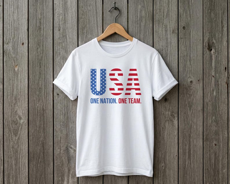 Peut inclure: Un t-shirt blanc &agrave; manches courtes avec le texte USA dans un motif bleu &eacute;toil&eacute; et ray&eacute; rouge et blanc. Sous les lettres, le texte ONE NATION. ONE TEAM. est imprim&eacute; en lettres capitales bleues et rouges sur le devant du v&ecirc;tement.