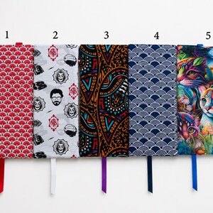 May include: Five rectangular fabric book sleeves arranged side-by-side. Pattern 1 is red with white fan shapes. Pattern 2 is white with black character faces and red logos. Pattern 3 is black with orange and blue geometric shapes. Pattern 4 is navy blue with white waves. Pattern 5 is a colorful cat print.