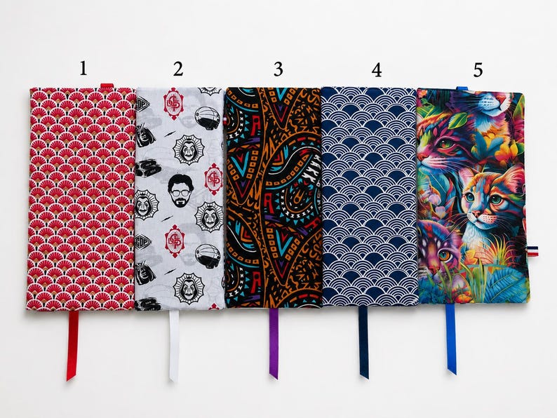 May include: Five rectangular fabric book sleeves arranged side-by-side. Pattern 1 is red with white fan shapes. Pattern 2 is white with black character faces and red logos. Pattern 3 is black with orange and blue geometric shapes. Pattern 4 is navy blue with white waves. Pattern 5 is a colorful cat print.