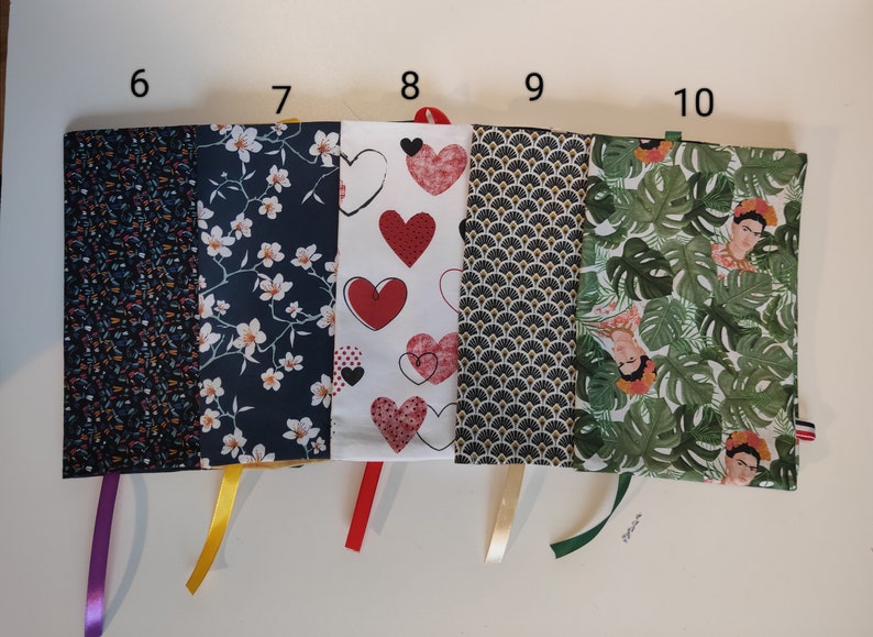 Puede incluir: Cinco fundas de libro de tela diferentes con lazos de cinta. Las fundas presentan varios patrones, incluyendo flores, corazones, geom&eacute;tricos y hojas tropicales con un retrato de Frida Kahlo.