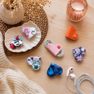 May include: A collection of heart-shaped cable organisers featuring various patterns and solid colours, including floral, geometric, and character prints. Each organiser is secured with a small snap button, designed to keep cords neatly bundled.