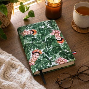 Può includere: Un taccuino rettangolare con un motivo ripetuto di ritratti di Frida Kahlo circondati da rigogliose foglie di monstera verdi. La copertina in tessuto ha uno sfondo bianco, un segnalibro a nastro nero e una piccola etichetta rossa, bianca e blu.