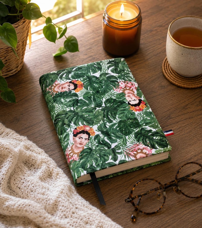 May include: A rectangular notebook featuring a repeating pattern of Frida Kahlo portraits surrounded by lush green monstera leaves. The fabric cover has a white background, a black ribbon bookmark, and a small red, white, and blue tag on the side.