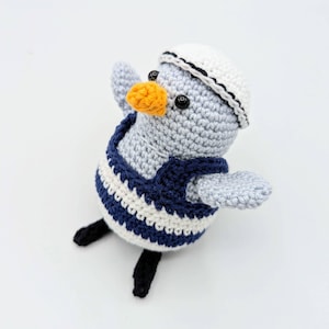 Peut inclure: Jouet de mouette crochetée vêtue d'une tenue de marin. La mouette est grise avec un chapeau de marin blanc, un gilet rayé bleu marine et blanc, et un bec orange. Le jouet a des yeux noirs en bouton et des pieds noirs.