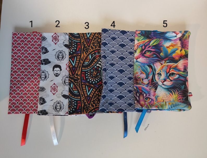 Puede incluir: Cinco cubiertas de libros diferentes con varios dise&ntilde;os. Las cubiertas est&aacute;n hechas de tela y tienen cintas de diferentes colores atadas a ellas. Los dise&ntilde;os incluyen un patr&oacute;n geom&eacute;trico rojo y blanco, un patr&oacute;n blanco y negro con la cara de un hombre, un patr&oacute;n paisley negro y naranja, un patr&oacute;n de olas azul y blanco y un patr&oacute;n de gato colorido.