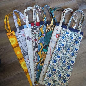 Puede incluir: Una colección de bolsas de mano coloridas con varios diseños, incluyendo girasoles, la Torre Eiffel, mariposas y diseños abstractos. Cada bolsa tiene un asa de tela a juego y un ribete en contraste.
