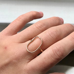 May include: A gold oval ring with a simple, open design. The ring is worn on a person's finger.