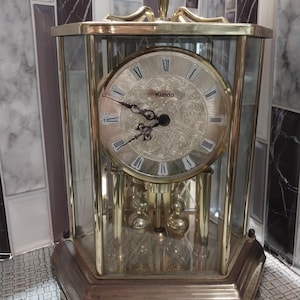 May include: A gold-toned Kundo brand clock with a glass case and a pendulum. The clock face has Roman numerals and a decorative design.