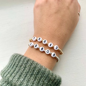 May include: Two beaded bracelets with white letter beads spelling out numbers. One bracelet has gold-colored beads and the other has silver-colored beads. The bracelets are worn on a wrist with a green sweater sleeve.