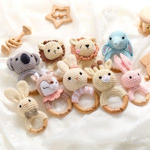 May include: A collection of eight crocheted animal baby rattles with wooden rings. The animals include a koala, two lions, a deer, a giraffe, two bunnies, and an elephant. Each rattle has a different name engraved on the ring, including Olivia, Nancy, and Bella.