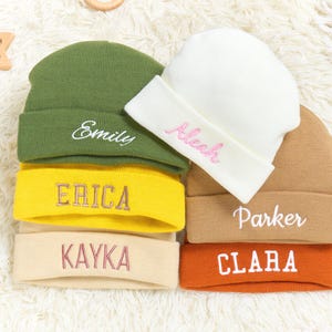 May include: Six beanies in a stack, in shades of green, yellow, beige, white, brown, and orange. Each beanie has a different name embroidered on the folded brim in a contrasting colour.
