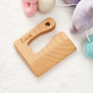 May include: A light brown wooden teether shaped like a U with a hole in the top. The teether is engraved with the name "Luca".