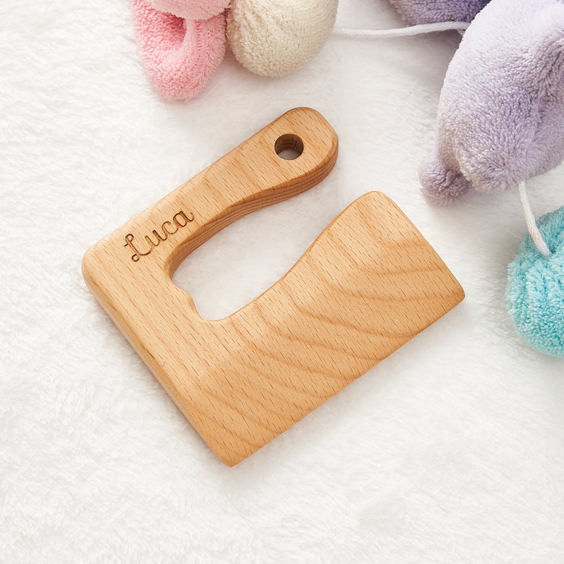 May include: A light brown wooden teether shaped like a U with a hole in the top. The teether is engraved with the name "Luca".