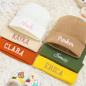 May include: Six personalised beanies in a stack, in shades of white, beige, orange, brown, green, and yellow. Each beanie has an embroidered name in a contrasting colour. The names are Aleah, Kayka, Clara, Parker, Emily, and Erica.