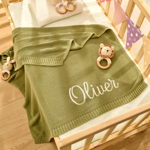 Puede incluir: Una manta de beb&eacute; de punto verde con el texto "Oliver" bordado en blanco. La manta est&aacute; doblada y colocada en una cuna de madera con un juguete de peluche de le&oacute;n y un anillo de madera.