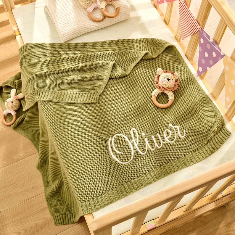 Puede incluir: Una manta de beb&eacute; de punto verde con el texto "Oliver" bordado en blanco. La manta est&aacute; doblada y colocada en una cuna de madera con un juguete de peluche de le&oacute;n y un anillo de madera.