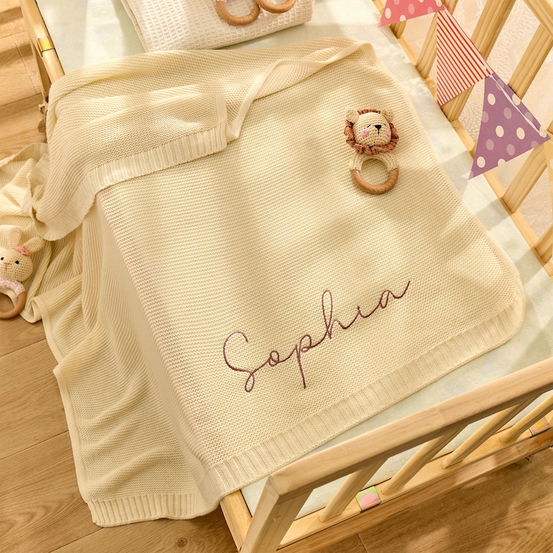 Puede incluir: Una manta de beb&eacute; de punto de color crema con el nombre "Sophia" bordado en rosa. La manta est&aacute; sobre una cuna de madera con un sonajero con forma de le&oacute;n.