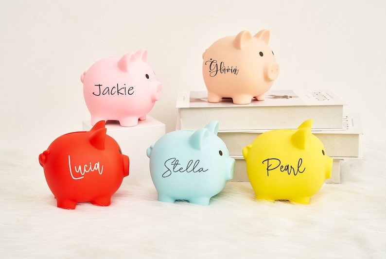 May include: Five piggy banks in different colors, each with a name written on them: Gloria, Jackie, Lucia, Stella, and Pearl.