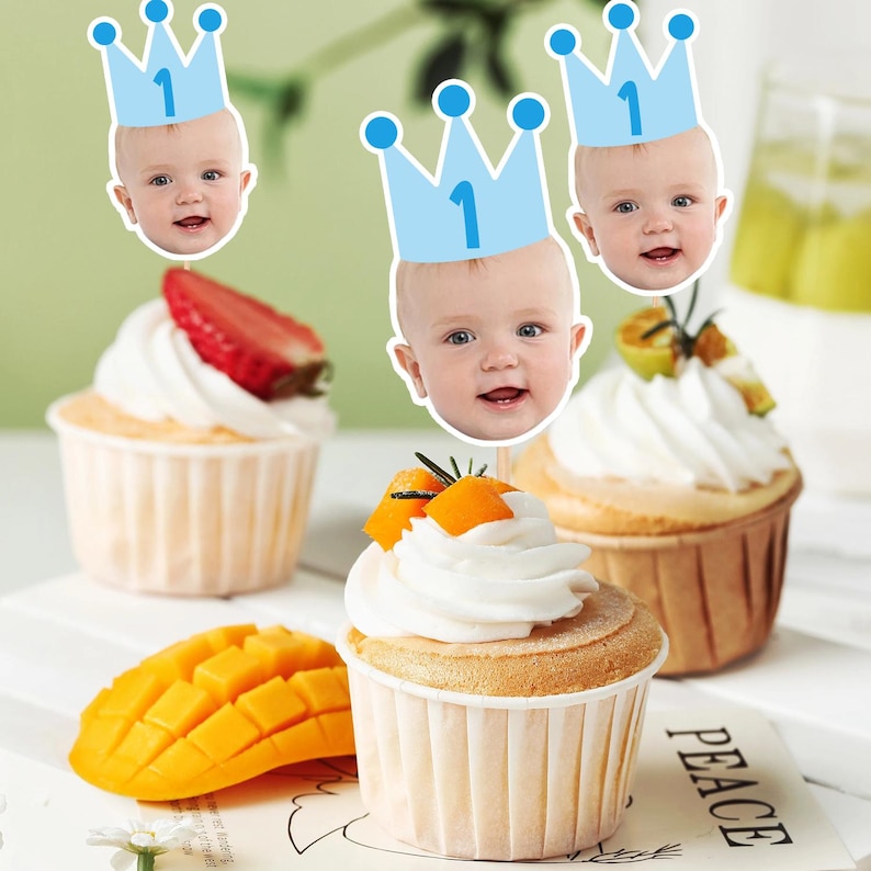 May include: Cupcakes with white icing, decorated with fruit and baby face toppers wearing blue crowns with the number 1. A mango slice is on the side. The cupcakes are in paper cases.