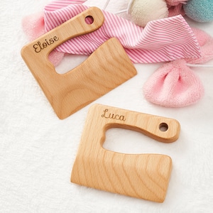 May include: Two wooden teethers with handles, one with the name "Eloise" engraved and the other with the name "Luca" engraved. The teethers are on a white fluffy surface.