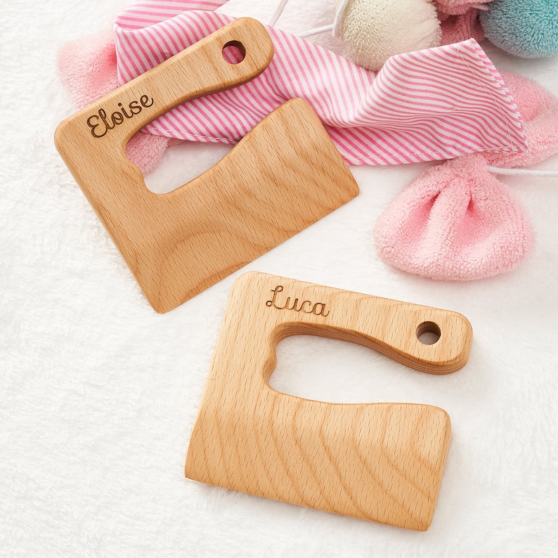 May include: Two wooden teethers with handles, one with the name "Eloise" engraved and the other with the name "Luca" engraved. The teethers are on a white fluffy surface.