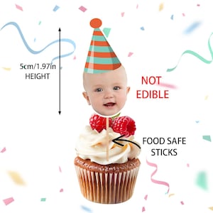 May include: A cupcake with white frosting, topped with raspberries and a baby face cake topper wearing a party hat. The topper is 5cm (1.97in) tall. Text reads "NOT EDIBLE" and "FOOD SAFE STICKS".