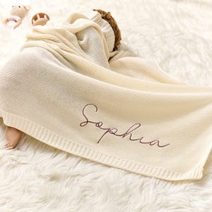 Puede incluir: Una manta de beb&eacute; de punto de color crema con el nombre "Sophia" bordado en color oro rosa.