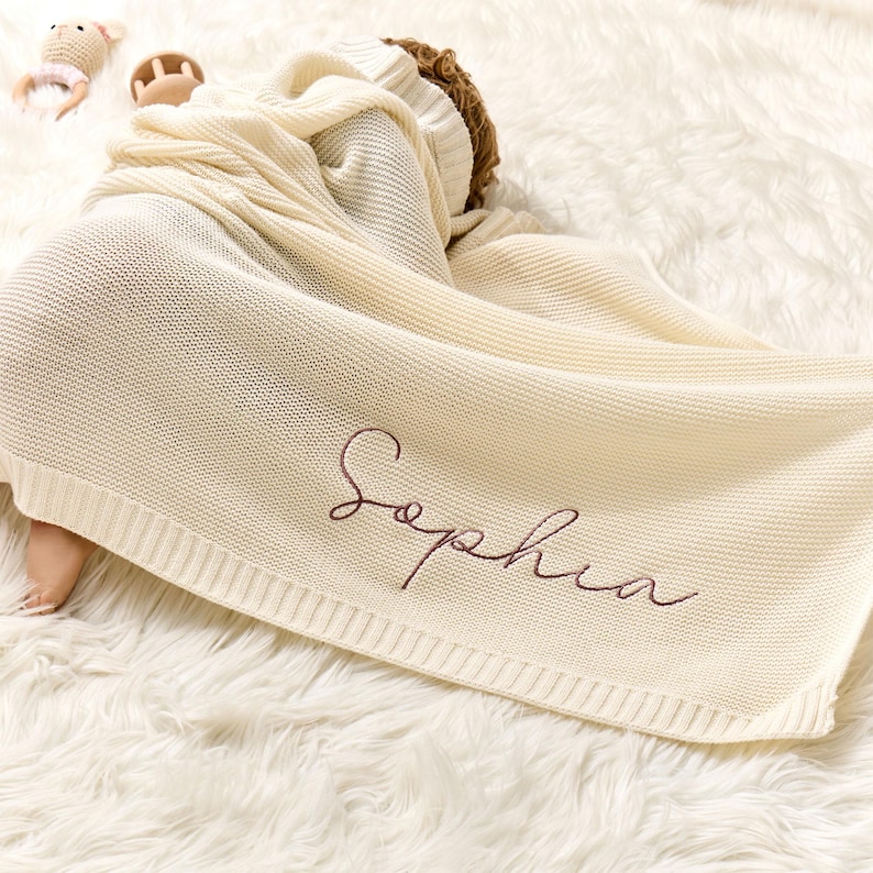 Puede incluir: Una manta de beb&eacute; de punto de color crema con el nombre "Sophia" bordado en color oro rosa.