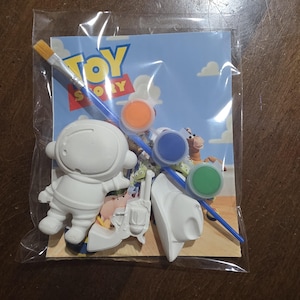 Könnte beinhalten: Ein klarer Plastikbeutel enthält ein Toy Story Malset. Das Set enthält eine weiße Astronautenfigur, einen Cowboyhut, eine Spielzeugpistole, drei Farbtöpfe, einen Pinsel und eine Karte mit dem Toy Story-Logo.