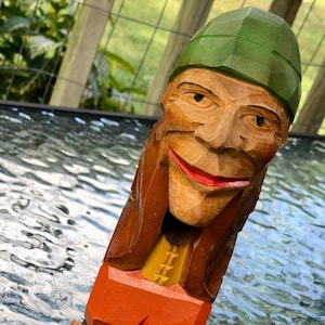May include: A wooden carved figurine of a person wearing a green hat, brown jacket, yellow shirt, orange pants, and black shoes. The figurine is standing on a wooden base.