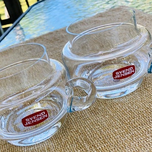 May include: Two clear glass mugs with handles. The mugs have a double-walled design and feature a red label with the text "SVEND JENSEN".