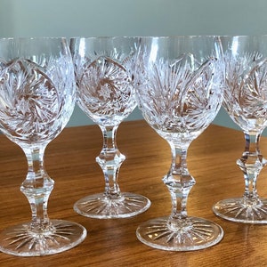 May include: Set of four clear crystal wine glasses with intricate cut designs. The glasses have a round bowl and a tall, slender stem.
