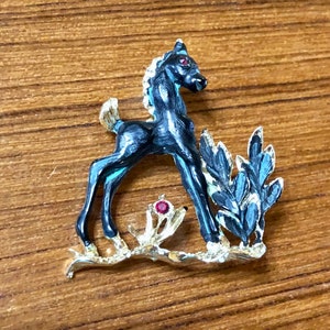 May include: A black enamel horse figurine with red eyes and gold accents standing on a gold branch with green leaves. The horse is a foal with a short mane and tail.