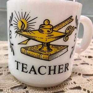 May include: White ceramic mug with a yellow and black illustration of an oil lamp, a book, and a quill pen. The mug says "TEACHER" in black lettering.