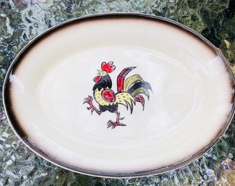 Puede incluir: Plato ovalado para servir con centro crema y borde marrón. En el centro, una colorida ilustración de un gallo con detalles rojos, amarillos, negros y verdes. El plato es probablemente de cerámica.