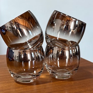 May include: A set of four rounded glass tumblers featuring a metallic bronze ombre finish that fades from a dark, reflective upper section to a clear, transparent base. The glasses have a smooth, curved silhouette and a wide, open rim design.