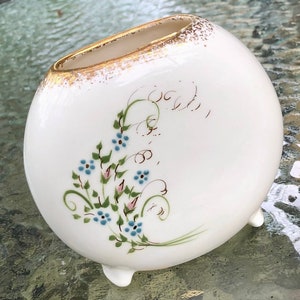 May include: White ceramic vase with a gold rim and floral design. The vase has three small feet and is decorated with blue and green flowers.